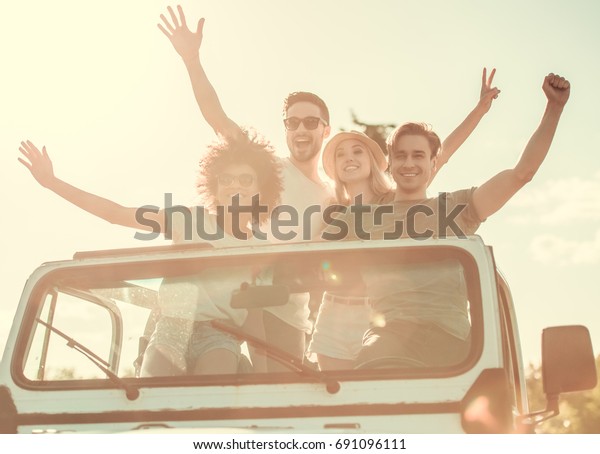 Happy young people are waving at\
camera and smiling while travelling by car in sunny\
weather
