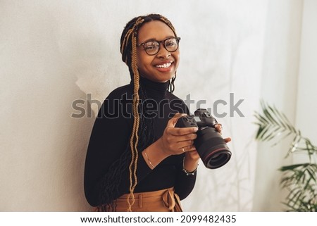 Happy young photographer smiling at the camera while standing against a wall in her home office. Creative female freelancer holding a dslr camera while preparing for a photo shoot.
