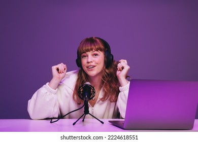 Happy young podcaster speaking into a microphone in a recording studio. Young woman hosting a live audio broadcast against a purple background. Woman creating content for her internet podcast. - Powered by Shutterstock