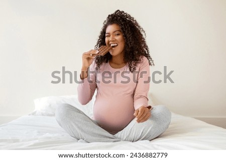 Happy young pregnant black woman enjoying chocolate bar while sitting cross-legged on white bed at home, cheerful african american expectant lady having sweet food cravings during pregnancy