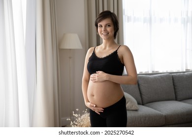 Happy Young Pregnant Woman In Black Underwear Holding Big Bare Baby Bump. Portrait Of Expectant Mother In Comfortable Bra Touching Smooth Belly Skin, Looking At Camera, Smiling. Skincare, Pregnancy