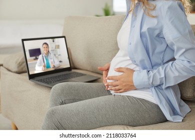 Happy Young Pregnant Woman Sitting On Sofa With Laptop, Holding Hands On Her Belly And Having Virtual Video Consultation With Online Doctor. Pregnancy, Expecting Baby, Health And Telemedicine Concept