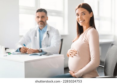 Happy young pregnant woman sitting at clinic during consultation with middle aged male gynecologist, embracing belly and smiling at camera, selective focus on lady - Powered by Shutterstock