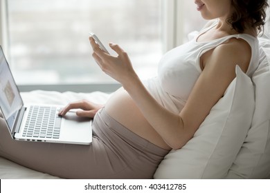 Happy young pregnant woman sitting on bed using technology devices: laptop and cellphone. Future mom expecting child relaxing at home bedroom, dialing number and looking at smartphone screen, close-up - Powered by Shutterstock