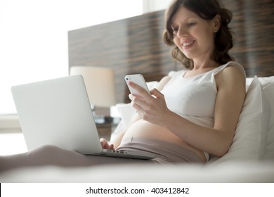 Fröhliche junge schwangere Frau, die mit technologischen Mitteln im Bett sitzt: Laptop und Mobiltelefon. Zukünftige Mütter erwarten, dass sich das Kind zu Hause mit dem Computer entspannt, die Telefonnummer gewählt und den Smartphone-Bildschirm betrachtet