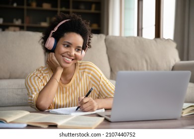 Happy young pretty African American woman in headphones looking at laptop screen, writing notes watching educational lecture, improving knowledge distantly, preparing for exams or studying remotely.