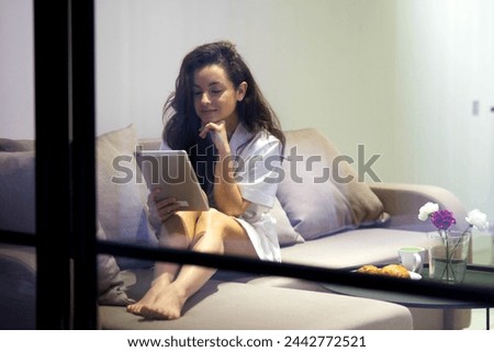 Happy young pretty Caucasian woman sitting on sofa in cozy apartment alone looking at gadget screen watching movie. Pretty brunette female indoor in living room resting using tablet device Stock fotó © 