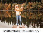 Happy young pretty woman walking outdoors in autumn park using mobile phone chatting, scenic view of the river