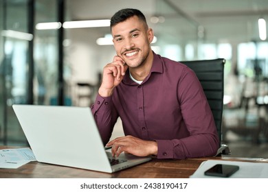 Happy young professional Latin business man looking at camera sitting at work desk using computer. Portrait of smiling male company worker employee, project manager or entrepreneur in office.