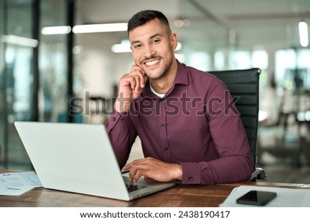 Happy young professional Latin business man looking at camera sitting at work desk using computer. Portrait of smiling male company worker employee, project manager or entrepreneur in office.