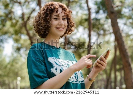 Happy young redhead woman standing on city park, outdoors hands holding cell phone touching finger display, scrolling on social media, mobile application or tech concepts. She is looking at the camera