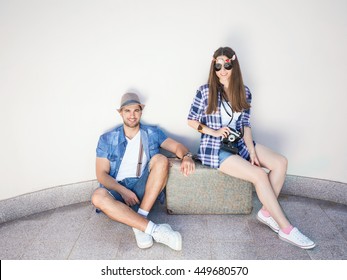 Happy Young Retro Styled Hippie Couple With An Old-fashioned Suitcase In Front Of The White Curved Wall. They Wear Matching Outfits. Young Man Is Sitting On The Floor And Looking At Camera