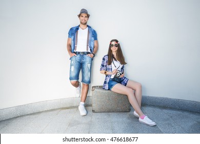 Happy Young Retro Styled Hippie Couple With An Old-fashioned Suitcase In Front Of The White Curved Wall. They Wear Matching Outfits.