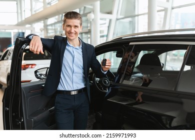 Happy Young Salesman Standing Near Open Car, Showing Auto Key, Selling New Vehicles At Modern Dealership, Empty Space. Handsome Millennial Manager Offering Choice Of Automobiles At Showroom Store