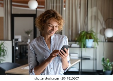 Happy young self employed woman using cellphone alone in office, reading text on screen, making mobile phone call, smiling, chatting online, getting good news, ordering, shopping via online app