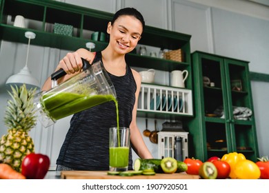 Happy young slim girl making fresh organic smoothie using blender in the kitchen