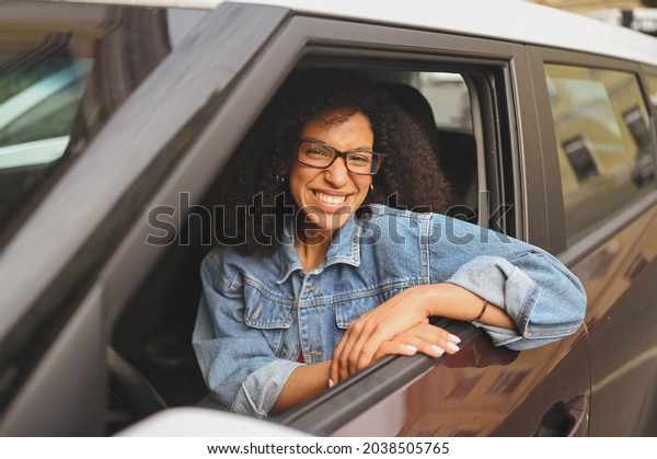Happy young smiling African American woman black\
haired driver in glasses sitting in new brown car, smiling looking\
at camera enjoying journey. Driving courses and life insurance\
concept.