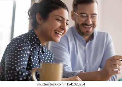 Happy young smiling diverse couple of female indian and male caucasian coworkers watching funny videos during coffee break, getting positive news, making good deal, holding video call with client