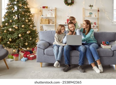 Happy young smiling family with two kids sitting on sofa using laptop together in festive living room with decorated Christmas tree enjoying winter holidays. New Year celebration concept. - Powered by Shutterstock