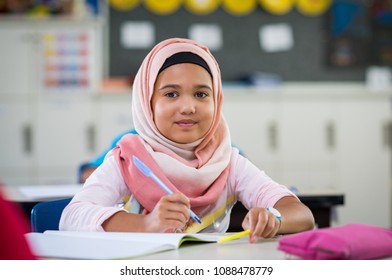 Happy Young Smiling Girl Wearing Hijab And Looking At Camera While Sitting At Desk With Exercise Book Elementary Muslim Schoolgirl Writing Notes In Classroom. Portrait Of Arab School Girl In Chador.