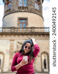 Happy young smiling Latin woman tourist in casual clothes and sunglasses using smartphone while standing against blurred historic Chapultepec Castle in Mexico City during trip