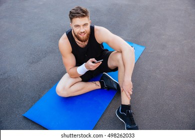 Happy young sportsman resting and using mobile phone with earphones while sitting on the blue mat outdoors - Powered by Shutterstock