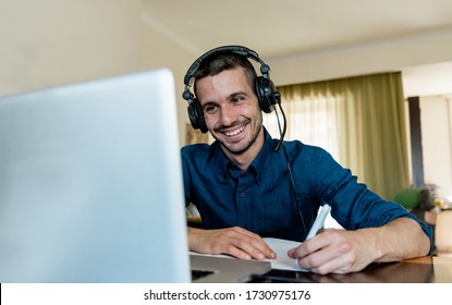 Happy Young Student At Home Studying On Computer Laptop Wearing Headphones Writing On A Book With A Pen. Online School Course Concept.