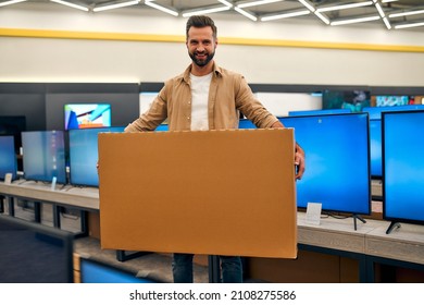 Happy Young Stylish Man Bought New TV, Holding Box With Purchase In Store Of Household Appliances, Electronics And Gadgets. Buying New TV, Sale Day.