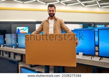 Happy young stylish man bought new TV, holding box with purchase in store of household appliances, electronics and gadgets. Buying new TV, sale day.