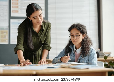 Happy young teacher of English language bending over desk with grammar tables illustrating usage of verb forms and tenses