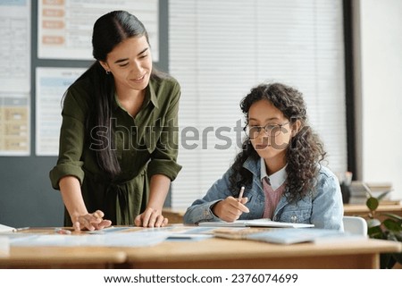 Happy young teacher of English language bending over desk with grammar tables illustrating usage of verb forms and tenses
