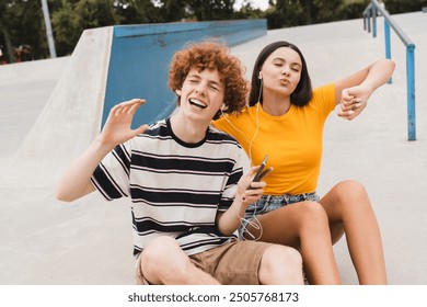 Happy young teenagers couple of college students high school pupils classmates friends listening enjoying music having fun sitting on skate park ramp spending time holidays together - Powered by Shutterstock