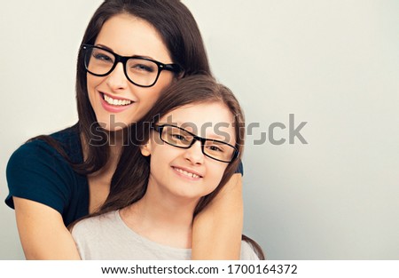Happy young toothy mother and smiling kid in fashion glasses hugging on light blue background with empty copy space for text. Closeup facial studio portrait