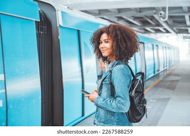 Happy young tourist African American woman with backpack and holding smartphone while at the train station and the train is arriving., Enjoying travel concept