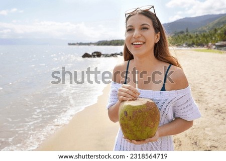 Happy young tourist woman holding green coconut on tropical beach. Summer vacation concept.