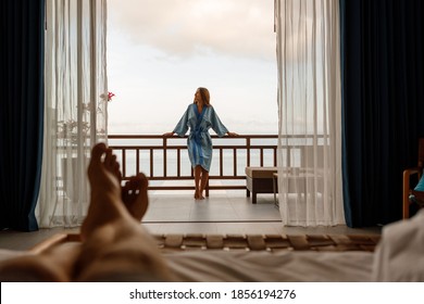 Happy young tourist woman stand on the balcony with sea view in hotel resort, wearing bathrobe. Man legs lie on the bead and look at girlfriend - Powered by Shutterstock
