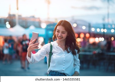 Happy Young Travel Asian Woman Using Mobile Phone And Selfie On Street Market Against Light Bokeh Background At Dusk In Bangkok, Thailand, Travel Vacation City Concept