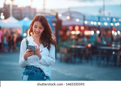 Happy young travel asian woman using mobile phone and relax on street market against light bokeh background at dusk in Bangkok, Thailand, Travel vacation city concept