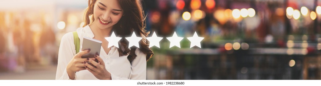 Happy young travel asian woman using mobile phone with graphic of five star icon to give rate of customer service mobile app on street market at dusk in Bangkok, Thailand - Shutterstock ID 1982238680
