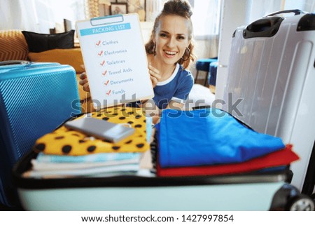 happy young traveller woman in blue t-shirt in the modern house in sunny summer day showing packing list.