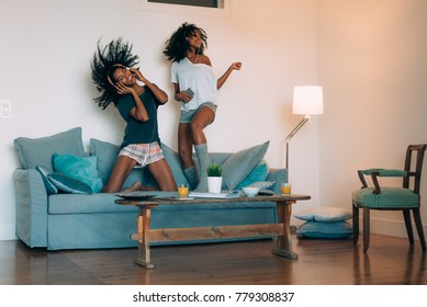 Happy Young Two Black Women Sitting And Standing Up In The Couch On The Computer And Mobile Phone With Headphones Dancing 
