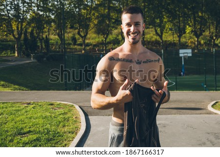 Happy young urban bodybuilder with tattoos taking off his shirt to to perform calisthenics exercises outdoors