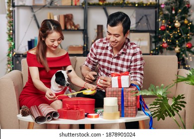 Happy young Vietnamese couple with cute small dog cutting colorful ribbons and wrapping presents for Christmas - Powered by Shutterstock