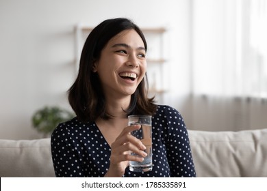 Happy young Vietnamese woman sit relax on sofa at home look in distance drink pure mineral water form glass. Smiling millennial Asian female enjoy clear still aqua. Healthy lifestyle concept. - Shutterstock ID 1785335891
