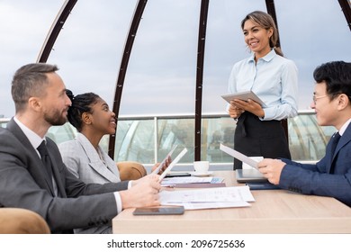 Happy Young Waitress With Tablet Taking Orders Of Intercultural Business Partners With Menus While Standing In Front Of Their Table