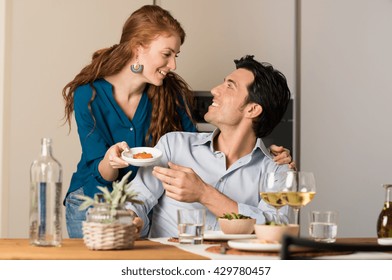 Happy Young Wife Serving Food To Her Husband. Romantic Couple Enjoying Lunch And Drinking Wine At Home. Young Woman Embracing Man During The Dinner.