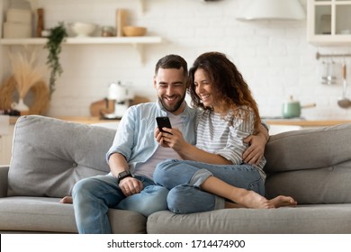 Happy young wife showing funny video in social network on cellphone to laughing husband. Smiling spouses shopping online web surfing, relaxing together on sofa in modern studio kitchen living room.
