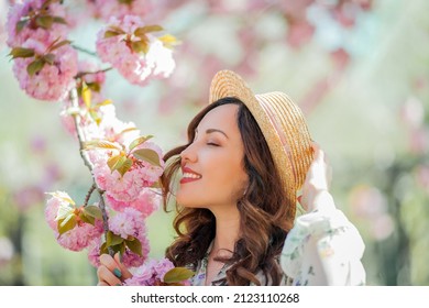 Happy young woman 30 years old in a wicker hat walks in a blooming city park. Portrait of a stylish emotional model in a light dress near pink flowering trees.