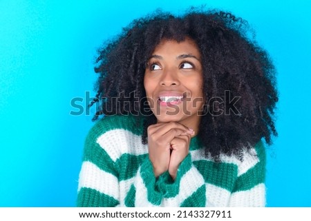 Happy Young woman with afro hairstyle wearing striped sweater over blue background anticipates something awesome happen, looks happily aside, keeps hands together near face, has glad expression.