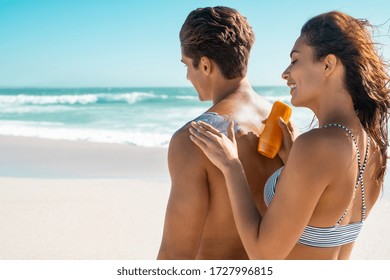 Happy young woman applying suntan lotion on boyfriend back at beach. Wife applying sunscreen lotion on man back at beach in sunshine. Girl put protection uv cream on shoulder of guy with copy space.